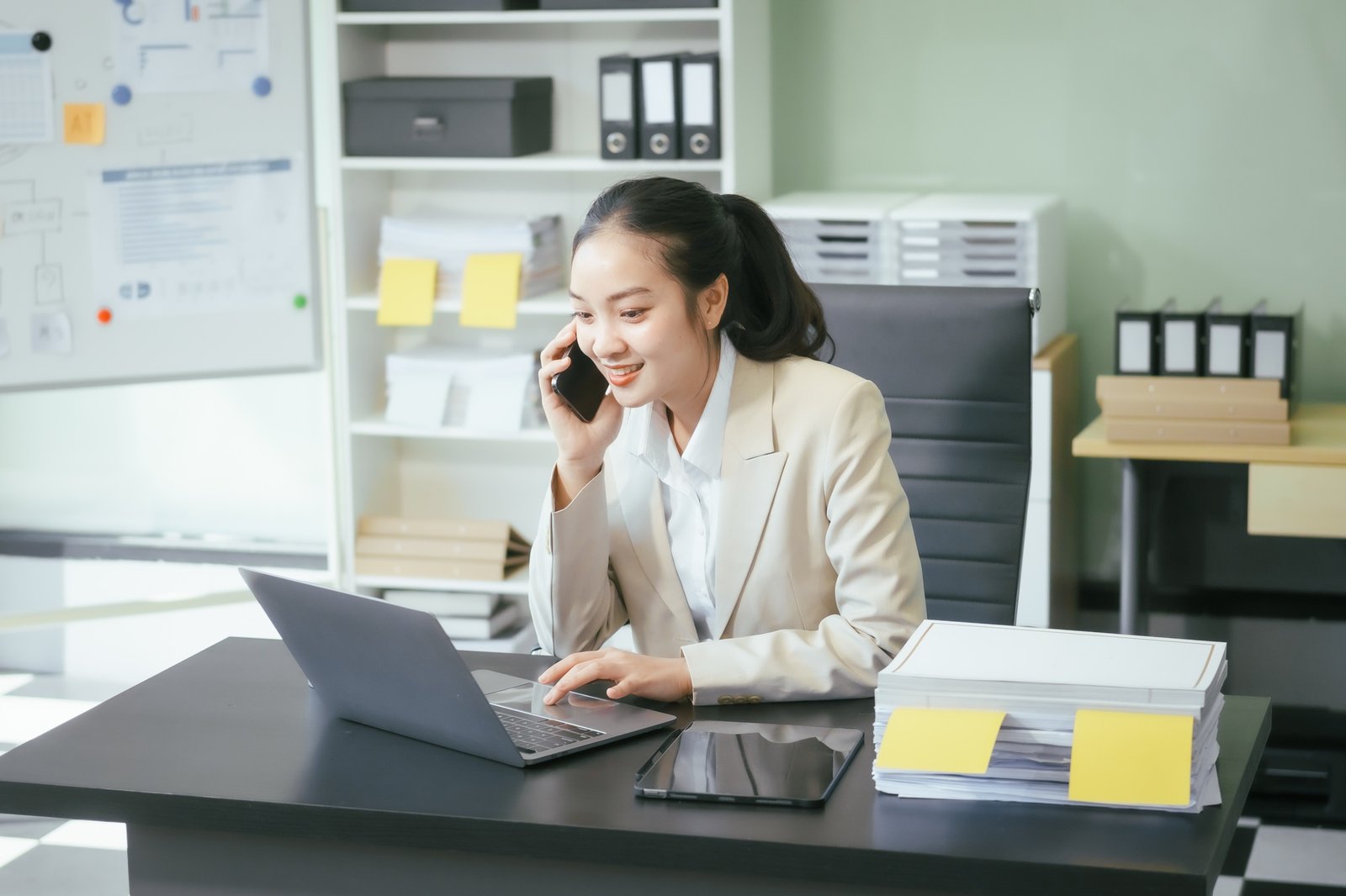 An Asian businesswoman sits at her office desk, smiling happily while working on her laptop